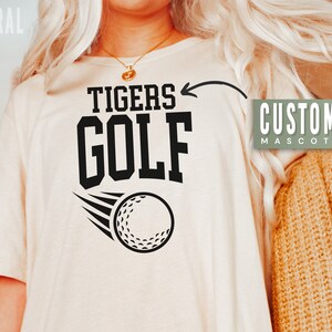 May include: A beige t-shirt with black text that says "Tigers Golf" and a golf ball graphic. The text "Customize Mascot" is in a green box on the right side of the image.
