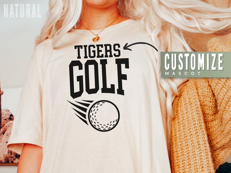 May include: A beige t-shirt with black text that says "Tigers Golf" and a golf ball graphic. The text "Customize Mascot" is in a green box on the right side of the image.