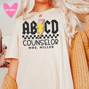 Pu&ograve; includere: Una maglietta bianca con testo nero che recita "ABCD Counselor Mrs. Miller". Il testo &egrave; in grassetto, in un carattere a blocchi. Un fulmine giallo con una gomma rosa in cima &egrave; al centro del testo.