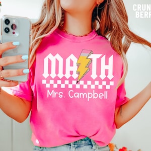 May include: Pink t-shirt with a white graphic that says "MATH" with a yellow lightning bolt and checkered pattern. The text "Mrs. Campbell" is below the graphic.