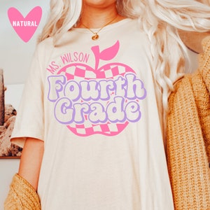 K&ouml;nnte beinhalten: Ein wei&szlig;es T-Shirt mit einem pinkfarbenen und violetten Apfeldesign. Der Apfel hat ein kariertes Muster und der Text "Ms. Wilson Fourth Grade" ist auf das Shirt gedruckt.
