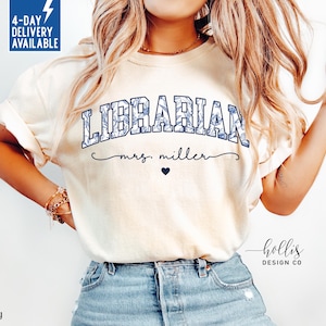 May include: Ivory-colored t-shirt with the word "LIBRARIAN" in a blue floral design. Below the word is "mrs. miller" in cursive, with a small blue heart. The shirt has a relaxed fit and is made of soft material.