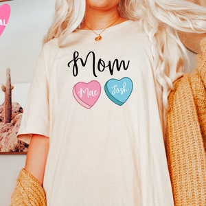 May include: White t-shirt with the word "Mom" printed in black cursive. Two candy hearts are below the word, one pink with "Mae" and one blue with "Josh" printed on them.