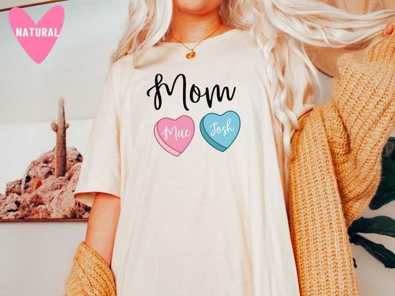 May include: White t-shirt with the word "Mom" printed in black cursive. Two candy hearts are below the word, one pink with "Mae" and one blue with "Josh" printed on them.