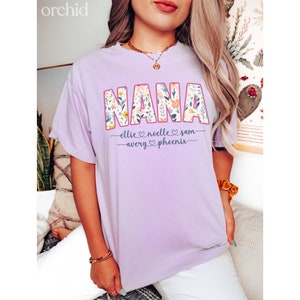 K&ouml;nnte beinhalten: Ein lavendelfarbenes T-Shirt mit dem Wort "NANA" in Blumenprint. Die Namen "Ellie, Noelle, Sam, Avery und Phoenix" stehen unter dem Wort "NANA" mit Herzen zwischen den Namen.
