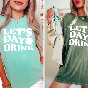 May include: Two women wearing green t-shirts with the text "Let's Day Drink" and a shamrock graphic. The t-shirt on the left is a light green color and the t-shirt on the right is a dark green color.