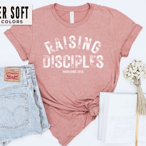 May include: A pink t-shirt with the text "Raising Disciples" in white, with the scripture reference "Proverbs 22:6" underneath. The shirt is made of a soft, comfortable material.