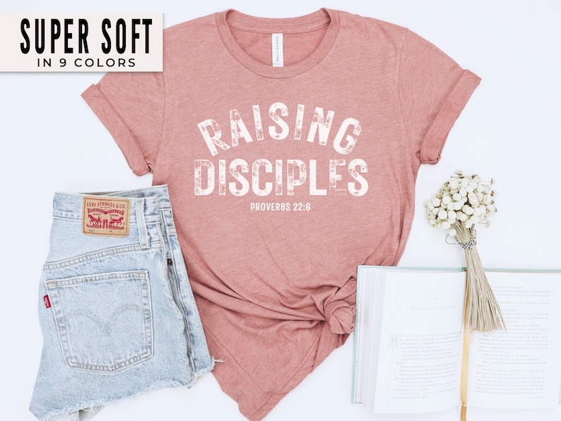 May include: A pink t-shirt with the text "Raising Disciples" in white, with the scripture reference "Proverbs 22:6" underneath. The shirt is made of a soft, comfortable material.