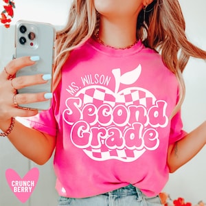 May include: Pink t-shirt with a white checkered apple design and the text "Ms. Wilson Second Grade".