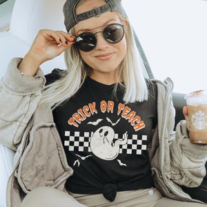Op de afbeelding: Een zwart T-shirt met een witte geestenafbeelding en de tekst "Trick or Teach" in oranje en zwart. Het shirt wordt gedragen door een persoon in een auto, die een kopje koffie vasthoudt.