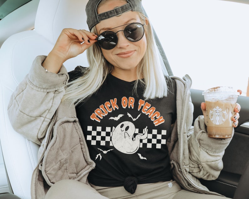 Op de afbeelding: Een zwart T-shirt met een witte geestenafbeelding en de tekst "Trick or Teach" in oranje en zwart. Het shirt wordt gedragen door een persoon in een auto, die een kopje koffie vasthoudt.