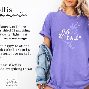 May include: A violet t-shirt featuring a white duck graphic and the words "dilly DALLY". The shirt has a casual fit and short sleeves. The image also displays the text "hollis guarantee".