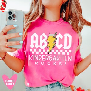 May include: Pink t-shirt with a white graphic that says "ABCD Kindergarten Rocks!" with a yellow lightning bolt and black and white checkered pattern.