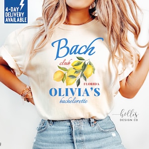 May include: Ivory-colored t-shirt with the text "Bach Club" in blue script, featuring a lemon and leaf graphic. The text "OLIVIA'S bachelorette FLORIDA" is also displayed in blue. The shirt has rolled-up sleeves.