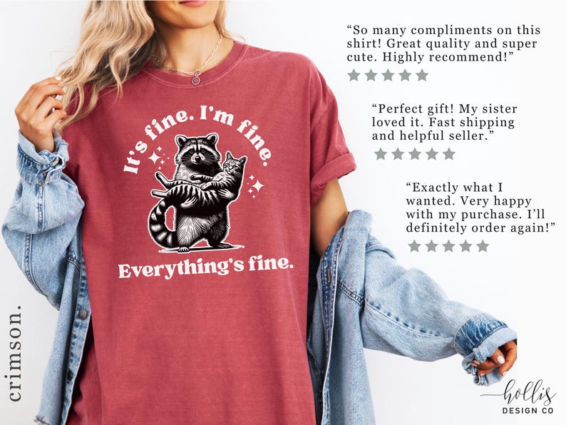 Puede incluir: Camiseta de color rojo carmes&iacute; con la frase "It's fine. I'm fine. Everything's fine." y un gr&aacute;fico de un mapache sosteniendo un gato. La camiseta tiene un ajuste c&oacute;modo y un aspecto desgastado.