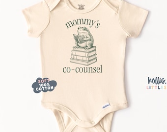 May include: Cream-colored baby onesie with short sleeves. The front features a green illustration of a frog reading a book atop a stack of books, with the text "mommy's co-counsel". A blue cloud graphic reads "SOFT 100% COTTON".