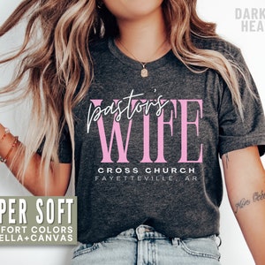 May include: A dark grey heather t-shirt with the text "pastor's wife" in pink lettering. The text "CROSS CHURCH FAYETTEVILLE, AR" is printed below the main text.