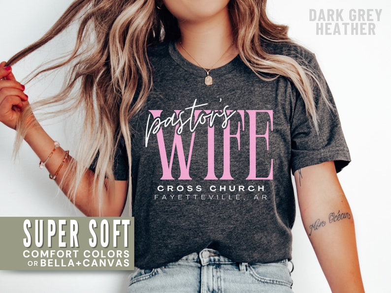 May include: A dark grey heather t-shirt with the text "pastor's wife" in pink lettering. The text "CROSS CHURCH FAYETTEVILLE, AR" is printed below the main text.