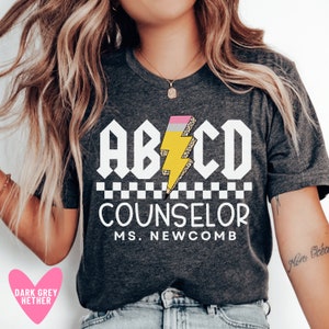 Pu&ograve; includere: Una maglietta grigia melange scuro con un design grafico bianco che dice "ABCD Counselor Ms. Newcomb". La grafica presenta un fulmine giallo con una gomma rosa e uno schema di stampa leopardata.