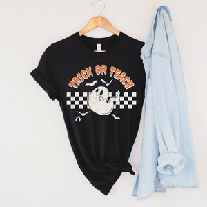 Op de afbeelding: Zwart T-shirt met een witte geest en de tekst "Trick or Teach" in oranje. De geest heeft een geruit patroon erachter en vleermuizen vliegen eromheen.
