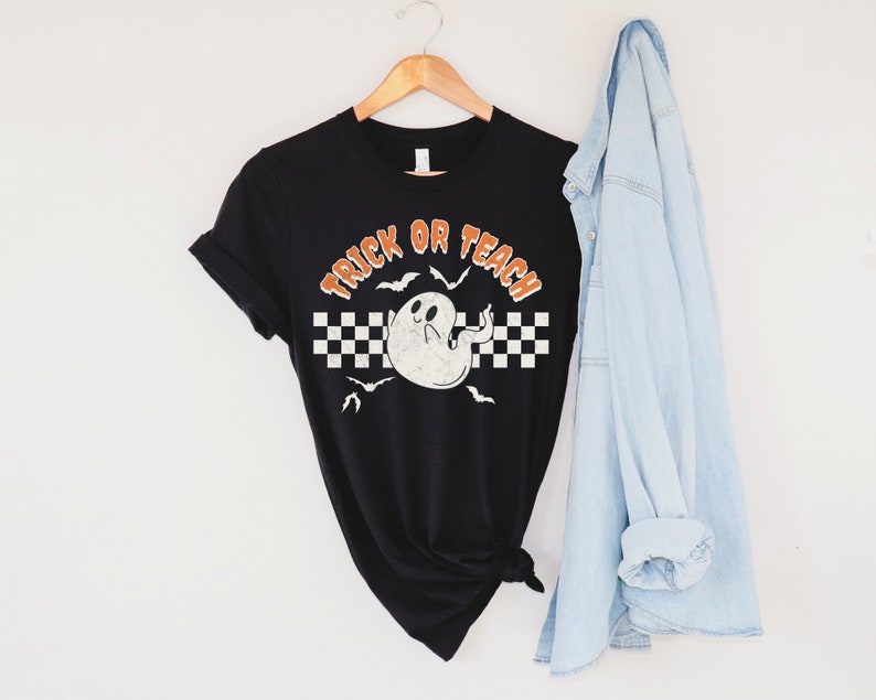 Op de afbeelding: Zwart T-shirt met een witte geest en de tekst "Trick or Teach" in oranje. De geest heeft een geruit patroon erachter en vleermuizen vliegen eromheen.