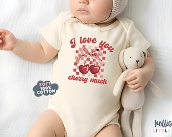 以下が含まれることがあります： 「I love you cherry much」の文字とチェリーのグラフィックが入った、クリーム色のベビーボディスーツ。柔らかい綿で作られており、市松模様が特徴です。赤ちゃんはウサギのぬいぐるみを持っています。