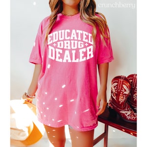 Op de afbeelding: Een oversized roze T-shirt met de tekst "EDUCATED + DRUG + DEALER" in witte letters.