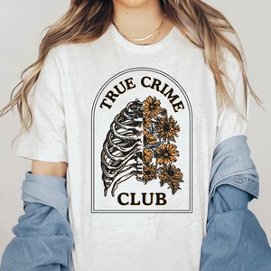 May include: White t-shirt with a graphic of a ribcage with flowers growing out of it. The graphic is surrounded by a black circle with the text "True Crime Club" in black and yellow lettering.