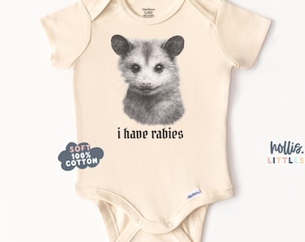 Pode incluir: Body de bebé creme com um gráfico de gambá a preto e branco e o texto "I have rabies". O body também tem um gráfico em forma de nuvem "Soft 100% Cotton". A marca "hollis.LITTLES" está impressa no lado direito.