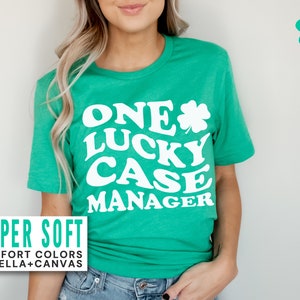 May include: A green t-shirt with the text "ONE LUCKY CASE MANAGER" in white, along with a white shamrock. The shirt also has the words "SUPER SOFT" and "COMFORT COLORS OR BELLA+CANVAS" in a white rectangle. The shirt is a crew neck.