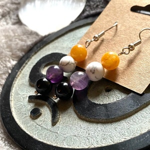 May include: A pair of dangle earrings featuring a combination of colorful gemstone beads. The earrings showcase a sequence of orange, white, purple, and black beads, suspended from silver-colored hooks. The earrings are displayed on a small, textured, decorative surface.