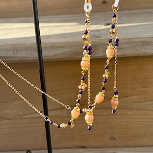 Può includere: Catene per occhiali color oro con perline color pesca, accenti viola e delicate catene dorate. Le catene sono esposte su un supporto in metallo nero, che mostra il loro design e i dettagli.