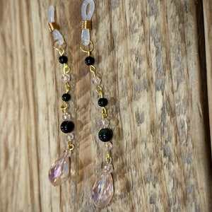 May include: Eyeglass chain with clear rubber loops, gold-colored metal, black beads, clear and pink crystal beads, and teardrop-shaped pink crystal pendants. The eyeglass chain is displayed against a wooden background.