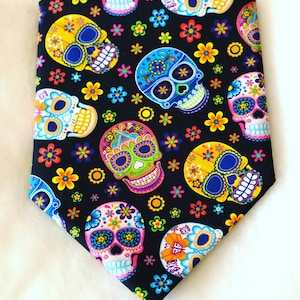 May include: A black fabric featuring a repeating pattern of colorful sugar skulls and flowers. The skulls are decorated with vibrant hues of blue, yellow, pink, and green, with floral accents. The flowers are in shades of orange, yellow, and blue.