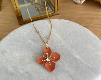 Könnte beinhalten: Eine goldfarbene Halskette mit einem Anhänger in Form einer korallenfarbenen Blume mit einem klaren Kristall in der Mitte. Die Halskette wird auf einer weißen Marmoroberfläche präsentiert, im Hintergrund eine goldgerahmte Glasbox.
