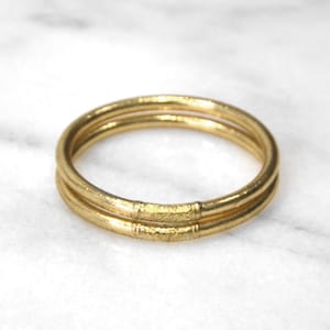 Peut inclure: Deux bracelets dorés empilés. Chaque bracelet a un design lisse et arrondi avec une bande texturée au milieu. Les bracelets sont présentés sur une surface en marbre blanc.