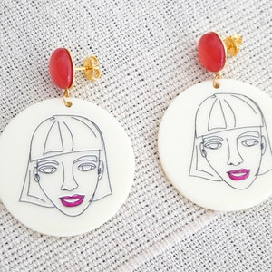 Peut inclure: Une paire de boucles d'oreilles circulaires blanches avec un dessin au trait d'un visage de femme sur chaque boucle d'oreille. Les boucles d'oreilles ont une pierre précieuse rouge attachée au sommet.