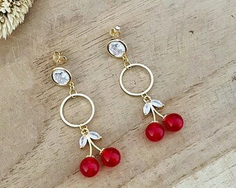Peut inclure: Boucles d'oreilles en forme de cerise dorées avec un strass transparent et un petit anneau doré. Les cerises sont rouges et ont une tige dorée avec de petites feuilles en strass.
