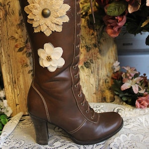 Könnte beinhalten: Braune Lederstiefel mit Absatz und Schnürung vorne, verziert mit drei Stoffblumen. Die Blumen sind weiß, beige und braun.