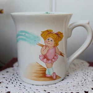 Op de afbeelding: Witte keramische mok met een handgeschilderde illustratie van een blond meisje in een roze tutu, dat een blauwe en groene toverstaf vasthoudt. Het meisje glimlacht en heeft staartjes.