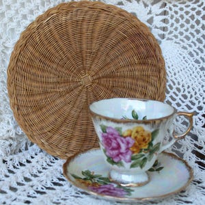 Könnte beinhalten: Ein Vintage-Tee-Tassen- und Untertassen-Set mit einem Blumendesign mit rosa und gelben Rosen, goldenen Verzierungen und einem gewebten Untersetzer aus Korbgeflecht. Die Tasse und Untertasse stehen vor einem weißen Spitzengrund.