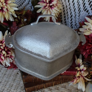 Könnte beinhalten: Eine silberne herzförmige Auflaufform mit Deckel und zwei Griffen. Die Form ist aus Gusseisen und hat eine strukturierte Oberfläche.