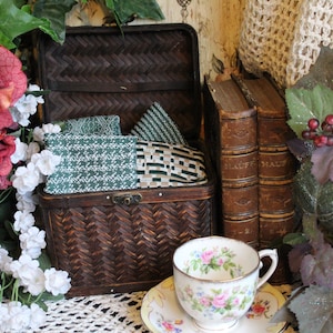 May include: A woven wicker basket with a hinged lid, filled with green and white patterned fabric squares. The basket is sitting on a white lace tablecloth next to two antique books and a teacup and saucer with floral designs.