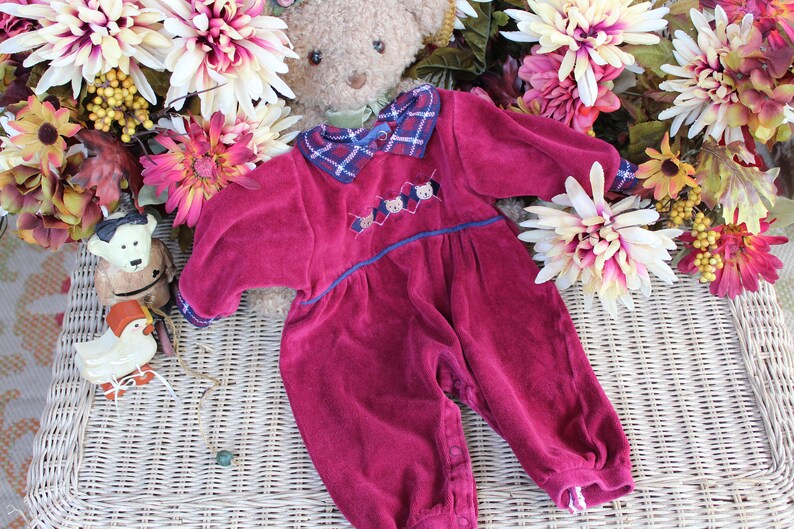 May include: A red velvet baby onesie with a plaid collar and embroidered bears. The onesie is on a wicker surface with flowers and a teddy bear.