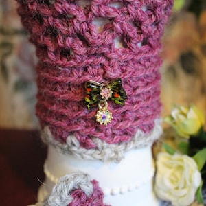 May include: A pink crocheted fabric sleeve with a decorative butterfly charm. The charm has a gold finish and features a red, white, and black design.