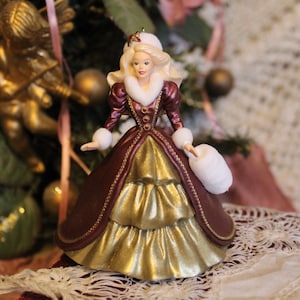 Op de afbeelding: Een porseleinen kerstversiering met een Barbiepop gekleed in een bordeauxrode en gouden jurk met een witte bontkraag. De pop houdt een witte bontmuff.