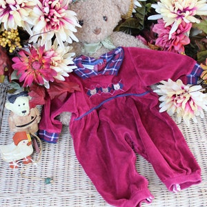 May include: A burgundy velour baby onesie with a plaid collar and embroidered bears. The onesie is lying on a wicker basket with fall-themed decorations.