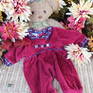 May include: A plush teddy bear wearing a burgundy corduroy jumpsuit with a plaid collar and embroidered bear faces. The bear is wearing a straw hat with a floral trim and is surrounded by fall flowers.
