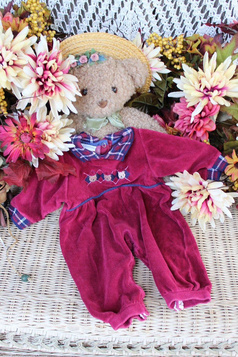 May include: A plush teddy bear wearing a burgundy corduroy jumpsuit with a plaid collar and embroidered bear faces. The bear is wearing a straw hat with a floral trim and is surrounded by fall flowers.