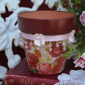 May include: A terracotta flower pot decorated with a pink ribbon, white bows, and crystal beads. The pot is decorated with a floral pattern featuring pink, red, and yellow roses.